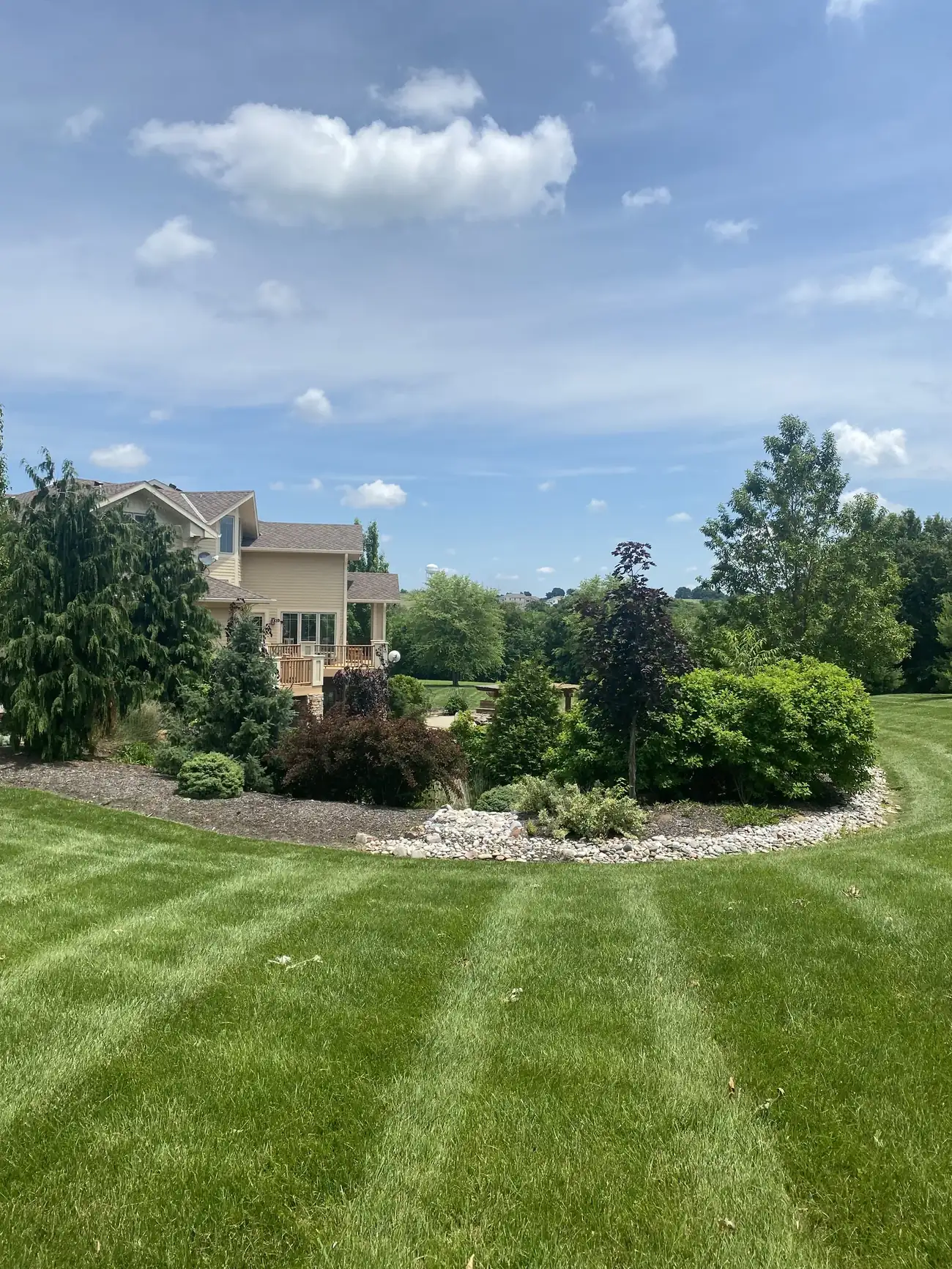 A landscaped bed with river rock and shrubs beside a striped lawn under a big spring sky